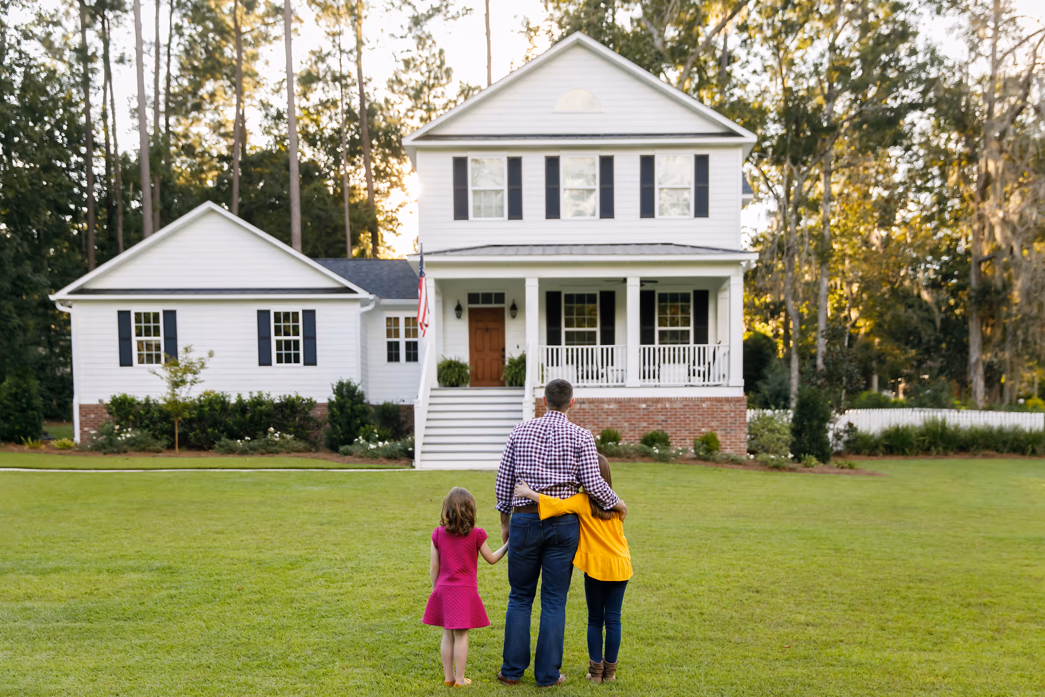 A parent and two children stand in a green front yard looking toward a well-maintained two-story white home with a covered porch and asphalt shingle roof.